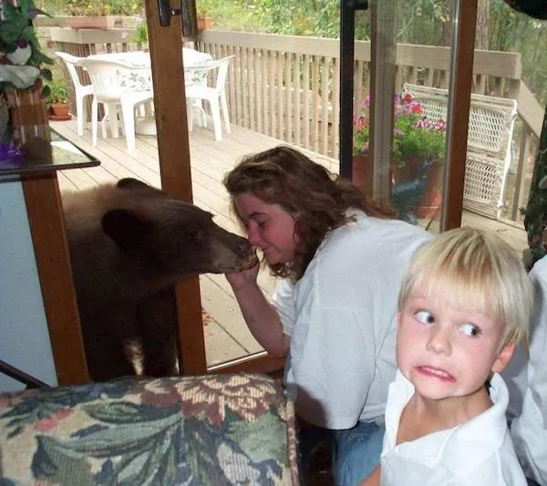 Zvedavý medveď na terase šnupá cez sklenené dvere, zatiaľ čo dieťa s vystrašeným výrazom pozoruje.