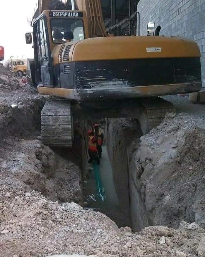 Báger nad výkopom s pracovníkmi kontrolujúcimi inštaláciu potrubia na stavbe.