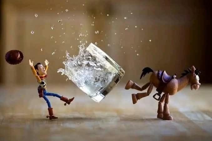 Hračka kovboja a koňa prevrhávajú pohár s vodou na drevenom pozadí, zaujímavá kompozícia pre zábavnú scénu.