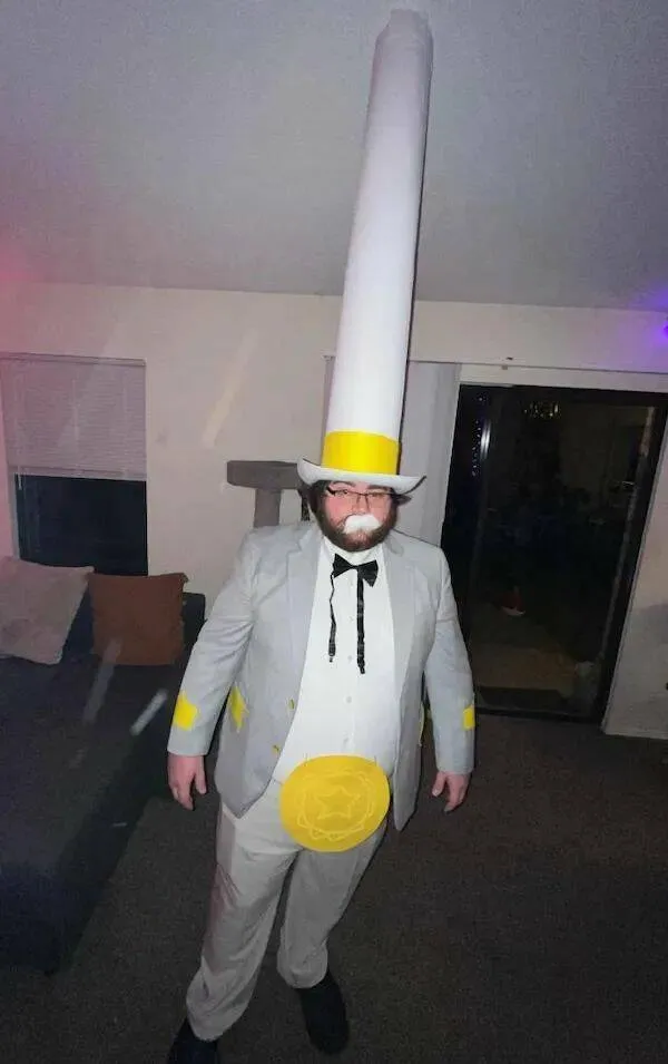 Osoba v kostýme cigarety s vysokým filtrom, šedý oblek, miestnosť s pohovkou a oknom v pozadí.