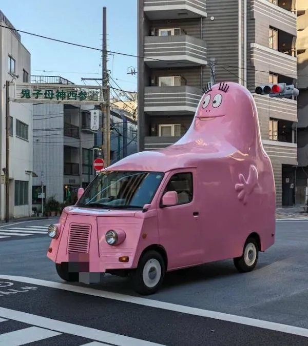 Ružové auto s kreslenou postavou na streche prechádza mestom, upútava pozornosť nápadným dizajnom.