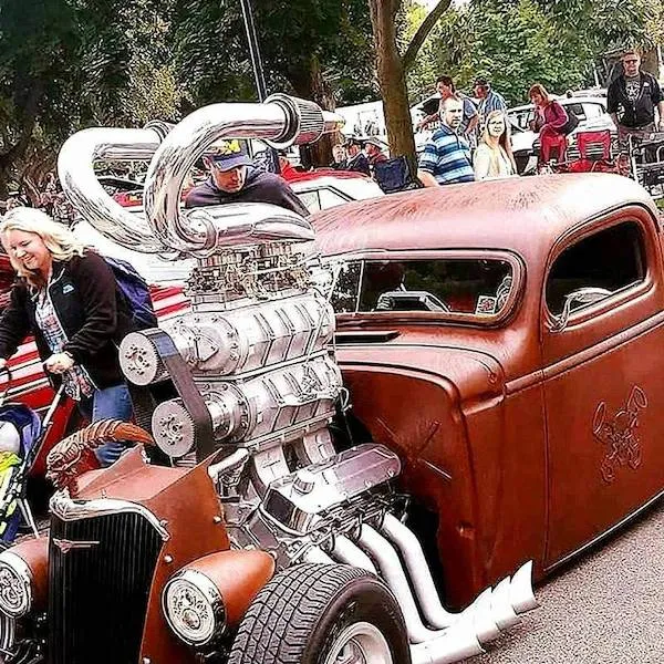 Staré auto s obrovským motorom vystavené na zraze veteránov, obklopené ľuďmi vonku na slnečnom dni.