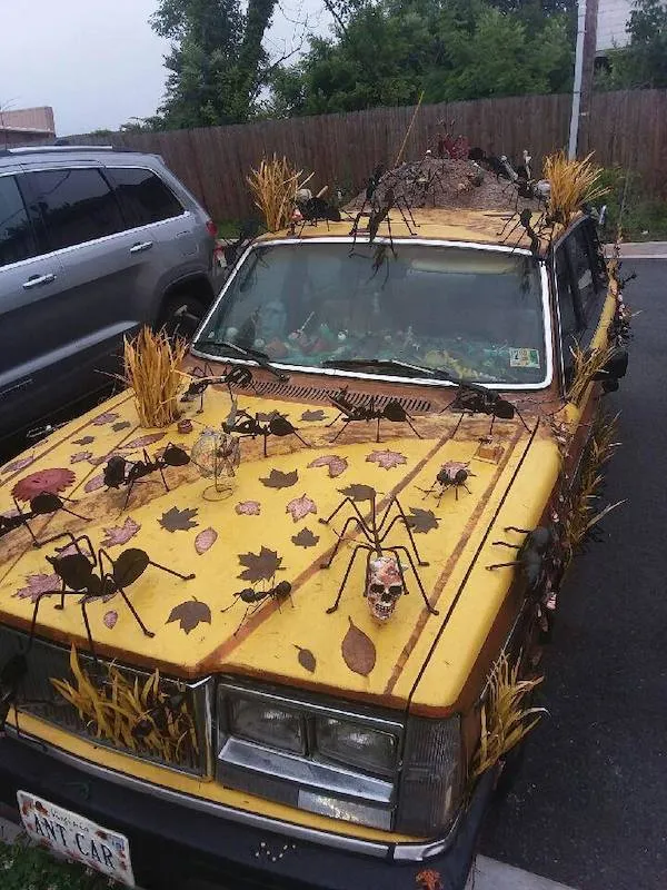 Auto pokryté umelými pavúkmi a listami, zaparkované vedľa SUV na asfaltovom parkovisku.