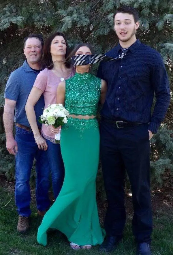 Ľudia pózujú na fotografii, jedna žena má tvár zakrytú kravatou vo vetre, zelené šaty a kvety v ruke.