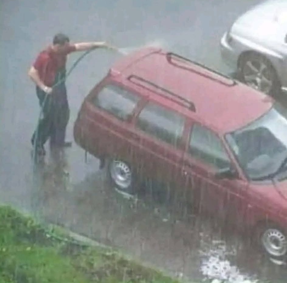 Muž umýva auto hadicou počas dažďa na ulici.