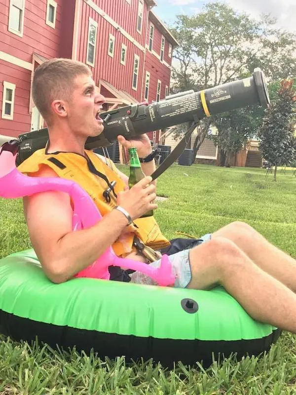 Muž v plavacej veste sedí na nafukovačke s fľašou a tubou na tráve pri červenom dome.