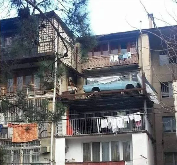 Auto zaparkované na balkóne panelového domu, pod ním sušiace sa prádlo, nezvyčajná situácia v mestskom prostredí.