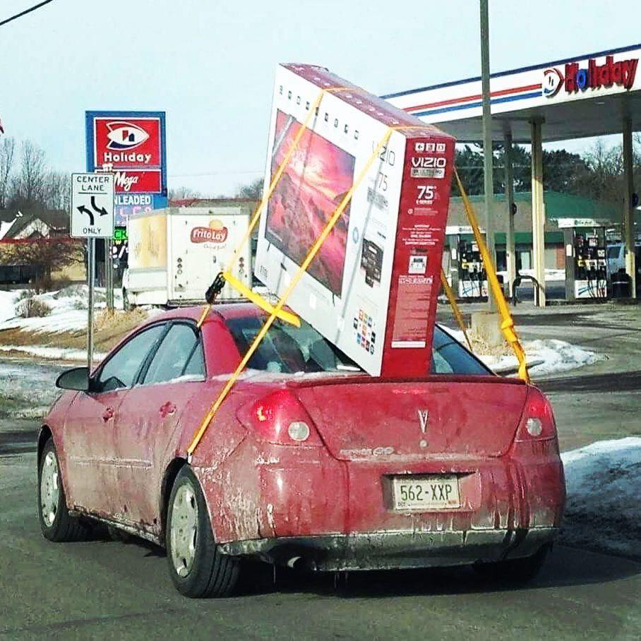 Červené auto s veľkou škatuľou s televízorom upevnenou na streche jazdí pri benzínovej pumpe počas zimy.