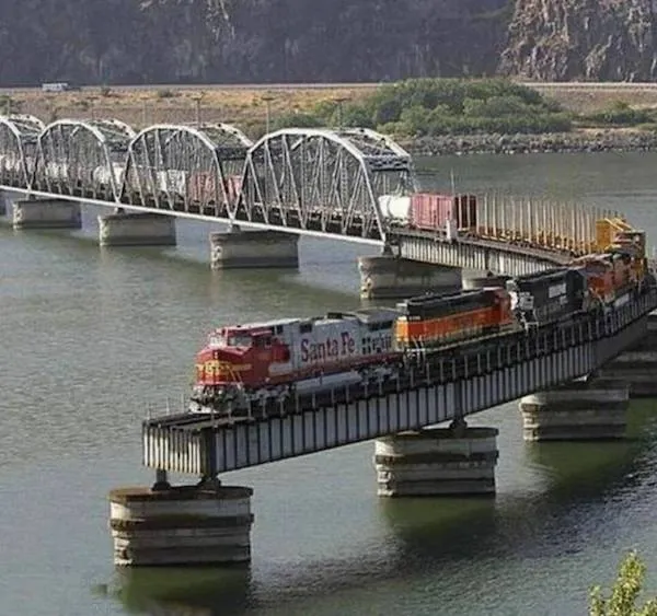 Vlak prechádzajúci cez riečny most s oblúkovou konštrukciou. Krajina s horami v pozadí.