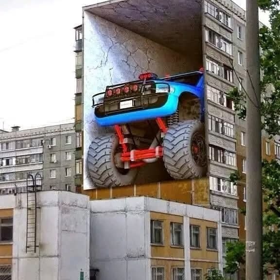 Nástenná maľba s veľkým modrým monster truckom na paneláku, realistický 3D efekt.