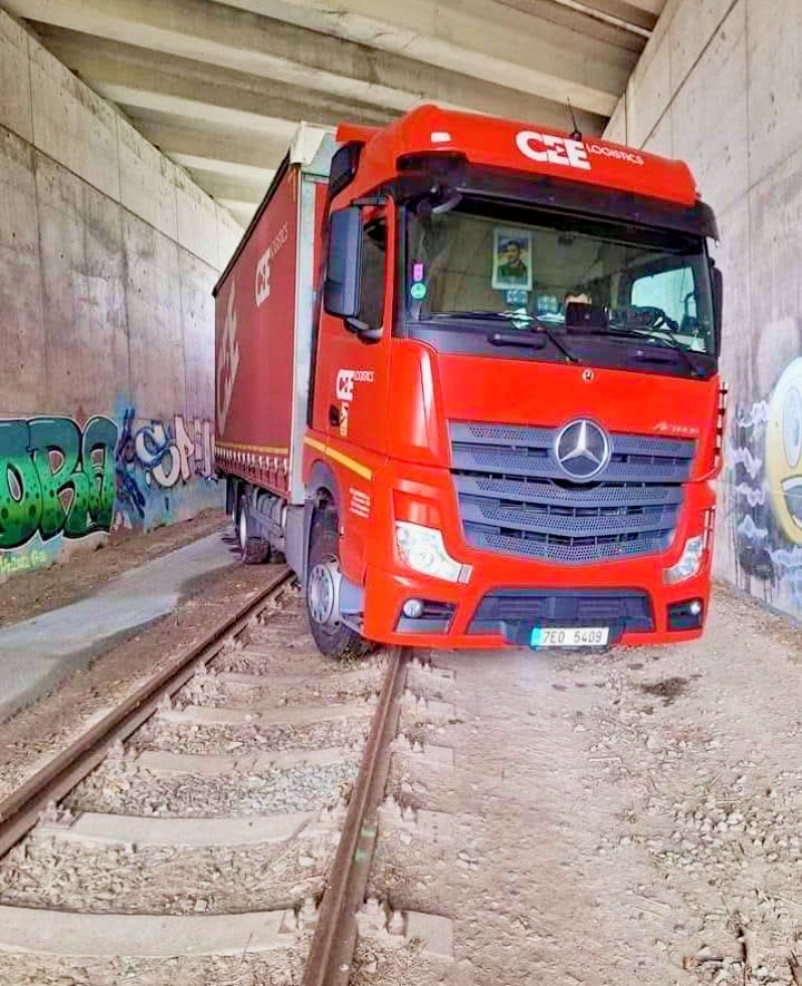 Červený nákladný automobil uviaznutý na železničnej trati pod mostom, obklopený graffiti na stenách.