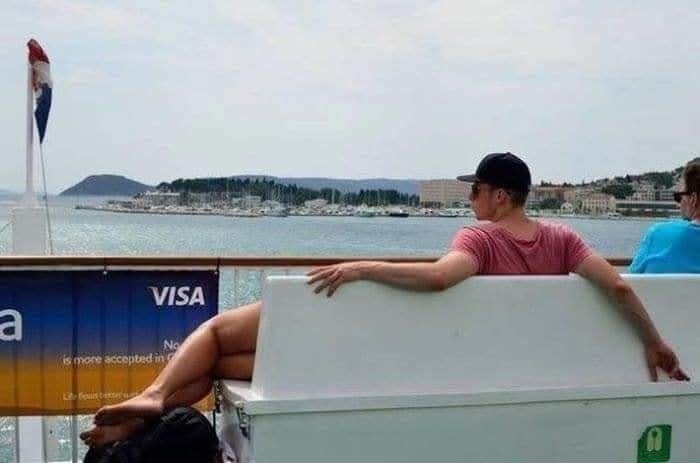 Muž sedí na lodi s výhľadom na more v pozadí, vedľa neho nohy druhej osoby, horúci letný deň.