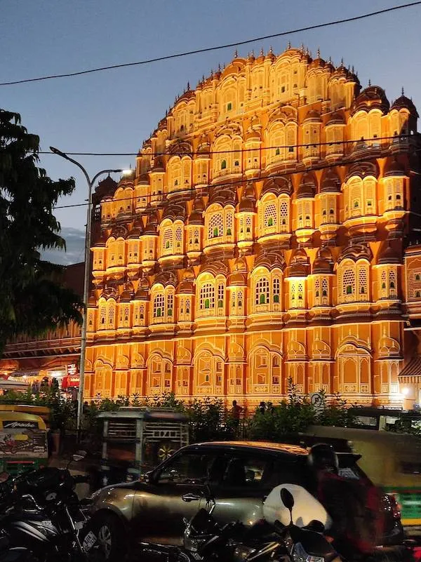 Osvetlená historická budova s komplikovanou architektúrou za súmraku, v popredí autá a skútre.