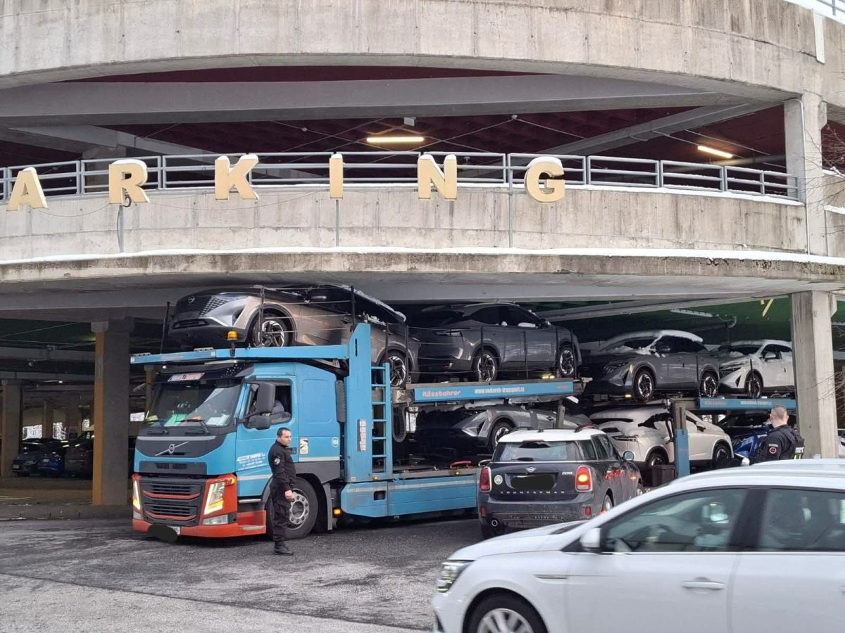 Kamión prevážajúci autá v parkovacom dome s nápisom „PARKING“, ľudia a autá v okolí.
