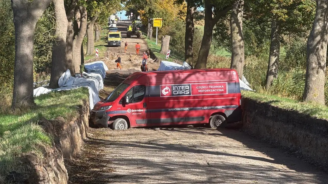 Červená dodávka uviaznutá naprieč cestou počas rekonštrukcie, robotníci pokračujú v stavebných prácach.