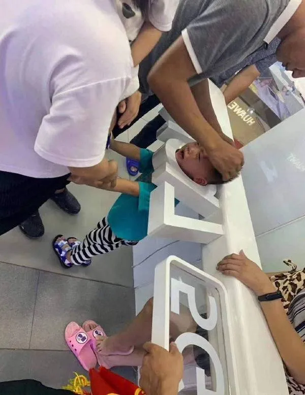 Dieťa s hlavou zaseknutou v medzere medzi pultom a písmenami, skupina dospelých sa snaží pomôcť.