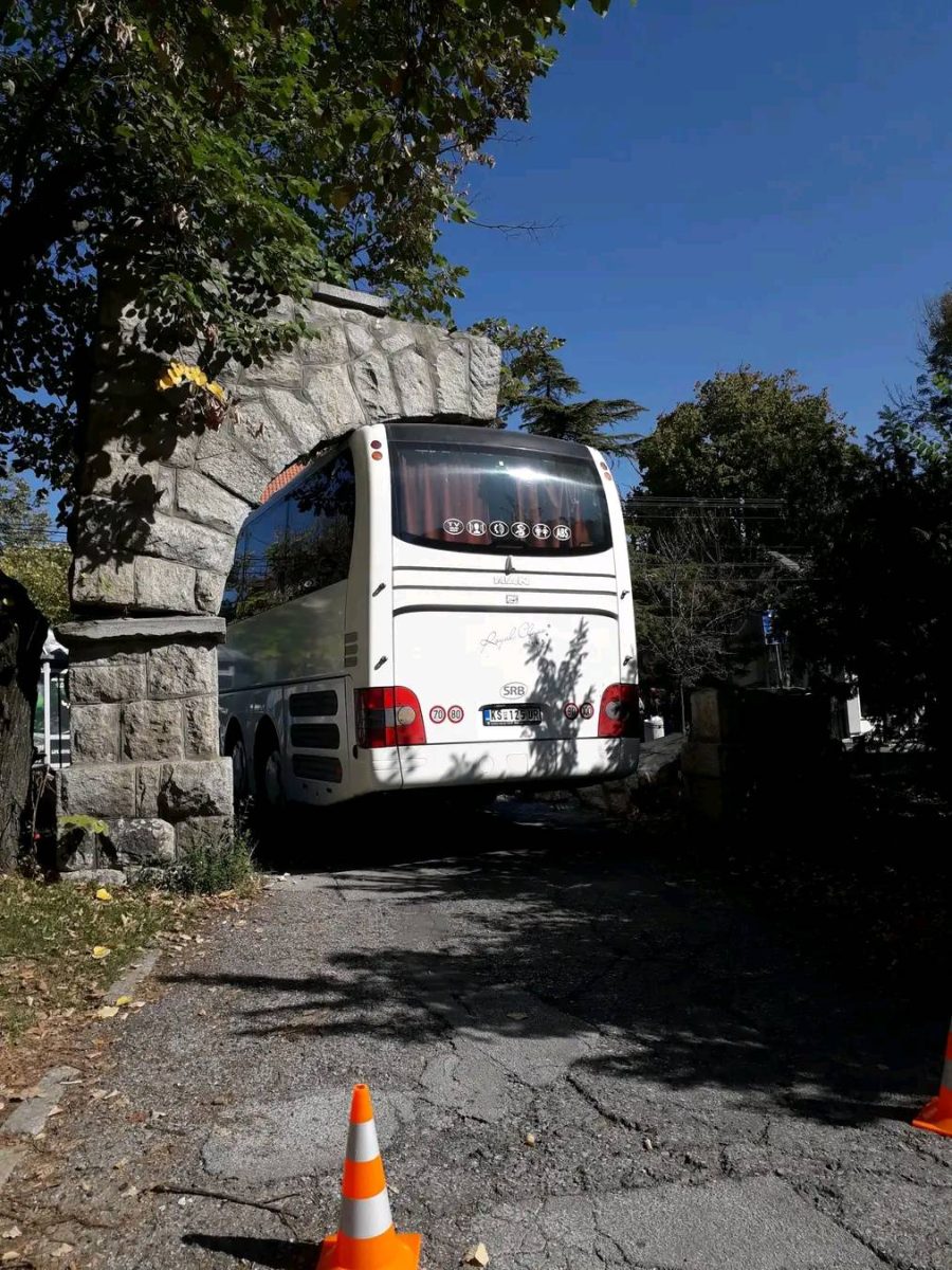 Autobus uviaznutý v kamennom oblúku na úzkej ceste, dopravné kužele pred ním, jasný deň s modrou oblohou.