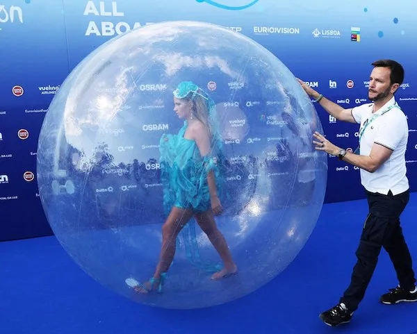 Osoba v modrom kostýme kráča vo veľkej priehľadnej guli na modrom koberci, modrá stena v pozadí.