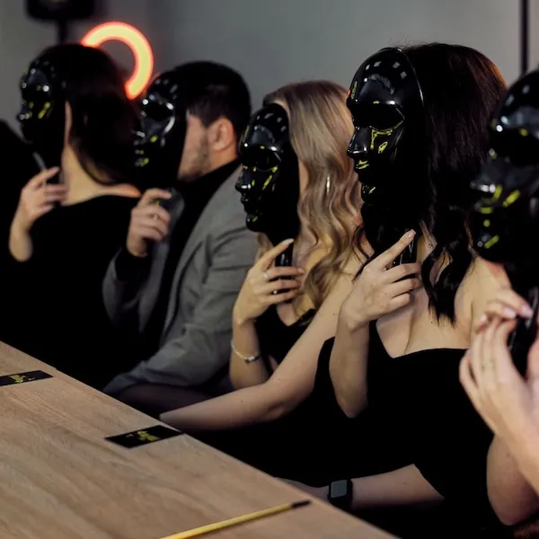 Skupina ľudí v čiernych maskách sedí pri stole počas záhadnej spoločenskej udalosti.