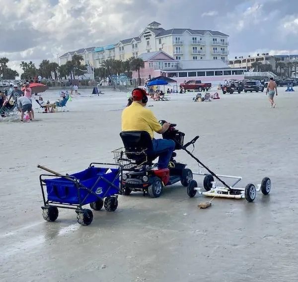 Osoba na elektrickom vozíku na pláži ťahá vozík s náčiním. Hotely a ľudia v pozadí.