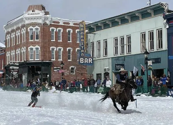 Lyžiar ťahaný koňom po zasneženej ulici pred historickou budovou, diváci sledujú jedinečnú zimnú udalosť.