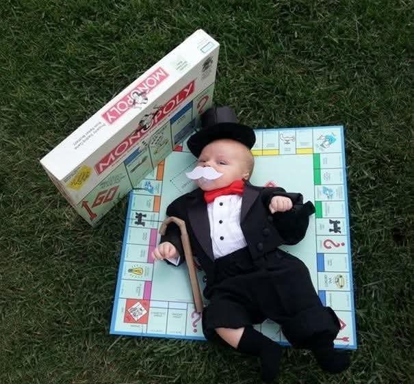 Dieťa oblečené ako Monopoly muž leží na hracej doske vedľa hernej krabice na tráve.