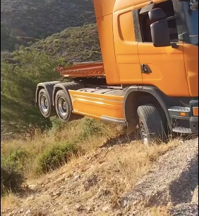 Oranžový kamión visí na okraji strmého svahu v horskej krajine.