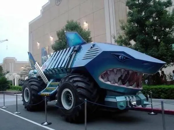 Obrovské auto v tvare žraloka s veľkými kolesami na parkovisku, obklopené zábranami.