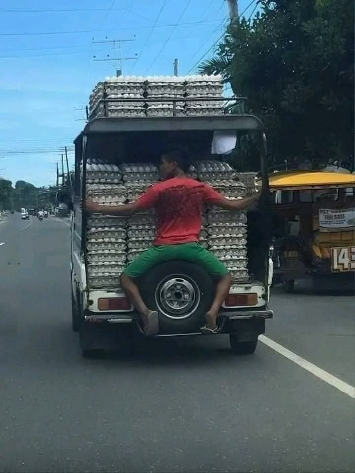 Muž sedí na korbe auta plného kartónov vajec, držiac sa za boku počas jazdy po ceste.