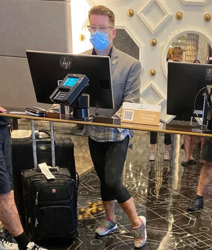 Muž s rúškom na recepcii hotela vedľa kufra, elektronická registrácia a informačná tabuľka.