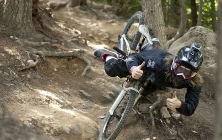 Cyklista v prilbe padá z bicykla na lesnej ceste, so zdvihnutými palcami hore.