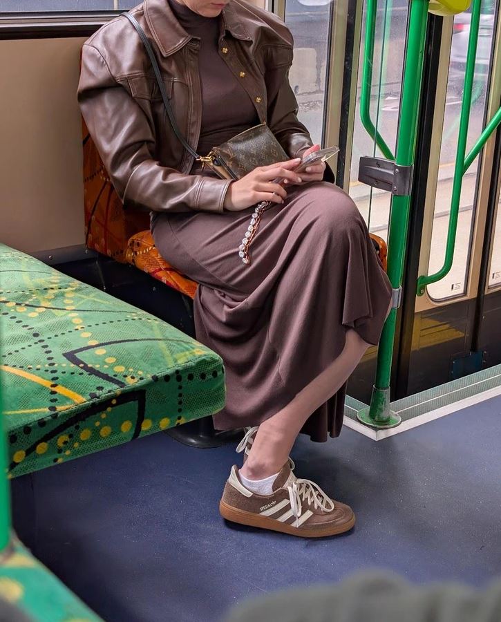Žena v hnedých šatách a bunde sedí v MHD, pozerá na mobil, obuté má športové tenisky a vedľa seba má kabelku.