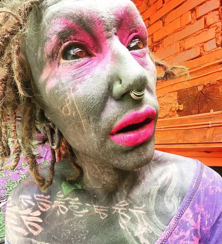 Osoba s farebným maľovaním na tvári a telu, s výraznými ružovými perami a piercingom v nose pred tehlovým múrom.