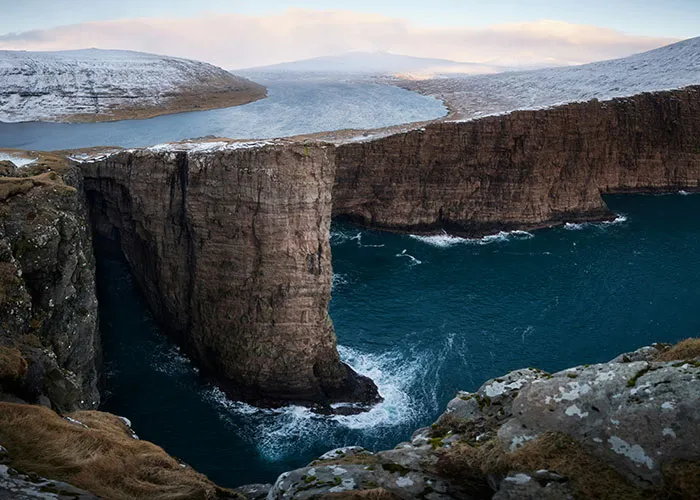 Skalný útes nad modrou vodou Severného Atlantiku na Farských ostrovoch, zasnežená krajina v pozadí.