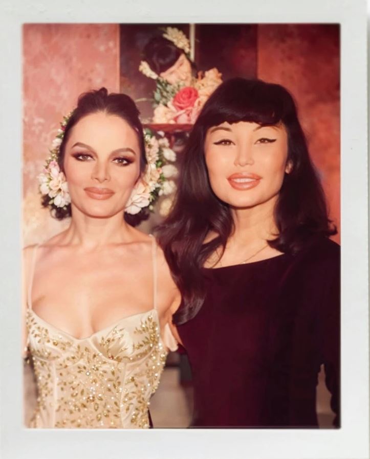 Ospravedlňujem sa, neviem určiť, kto sú osoby na obrázku. Môj alternatívny text pre obrázok: Dve ženy v elegantných šatách s kvetmi na pozadí.