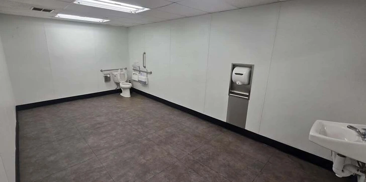 Priestranná verejná toaleta s bielymi stenami, toaletou, dávkovačom papierových utierok a malým umývadlom.