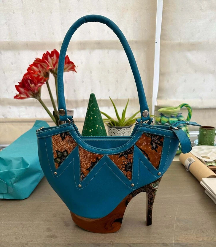 Modrá kabelka v tvare topánky s pestrofarebnými vzormi, umiestnená na stole vedľa rastlín a darčekového balenia.