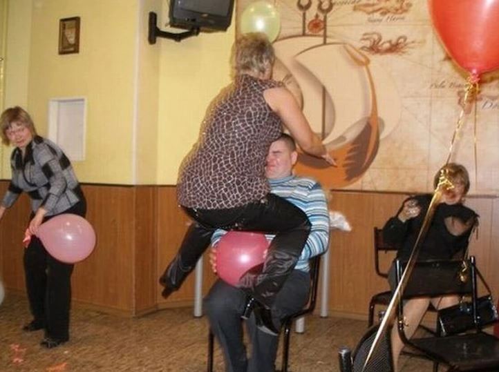 Na oslave sa ľudia bavia s balónmi, žena skáče na muža sediaceho na stoličke. Ostatní pozorujú a smejú sa.