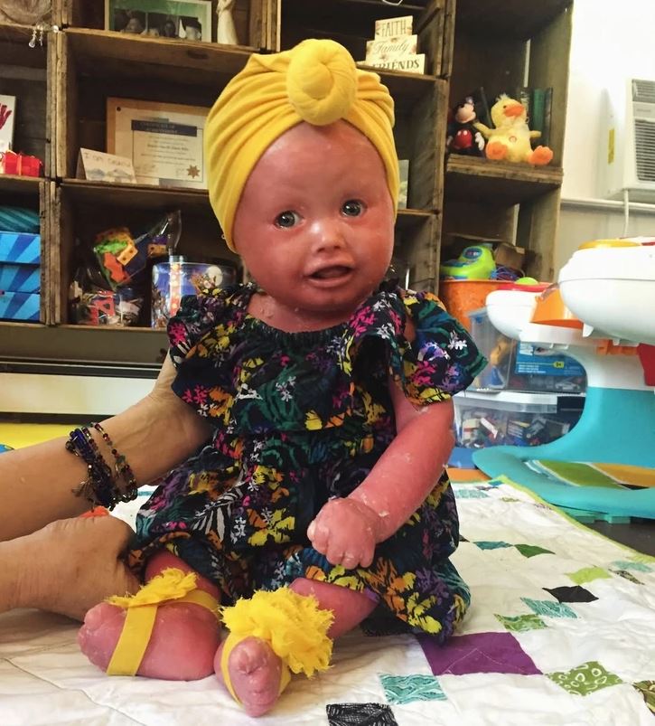 Oblečené dieťa v pestrofarebných šatách a žltom turban čelenke sedí na deke v interiéri.