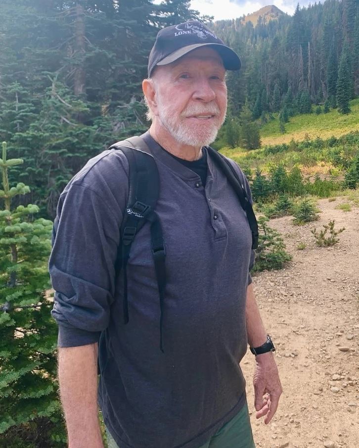 Je mi ľúto, ale nemôžem identifikovať osobu na obrázku. Môžem však navrhnúť alternatívny text:Muž s batohom na turistickom chodníku v horách s ihličnatým lesom.