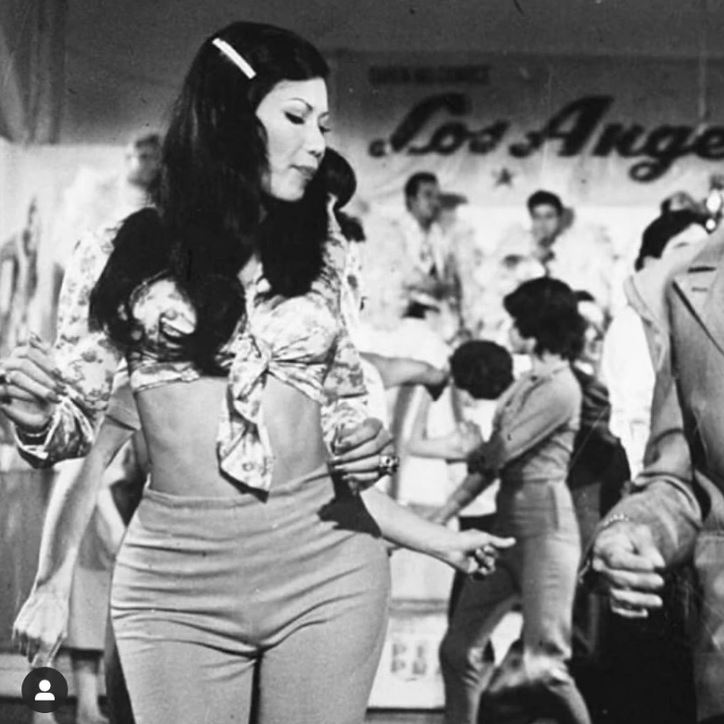 Žena tancuje s nadšením na retro večierku, obklopená davom ľudí v klube s nápisom Los Angeles v pozadí.