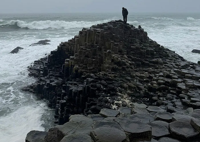 Osoba stojí na čadičových stĺpoch Giant's Causeway, obklopená vlnami oceánu pod zamračenou oblohou.