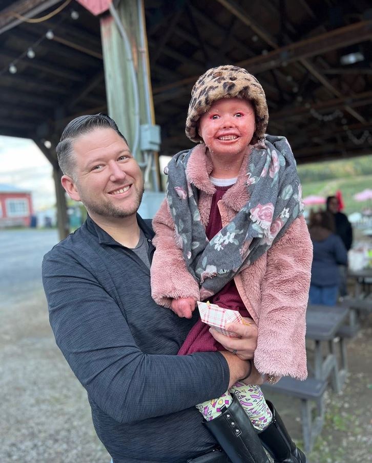 Otec drží usmievajúce sa dieťa v zimnom oblečení pod prístreškom na dvore s posedením.