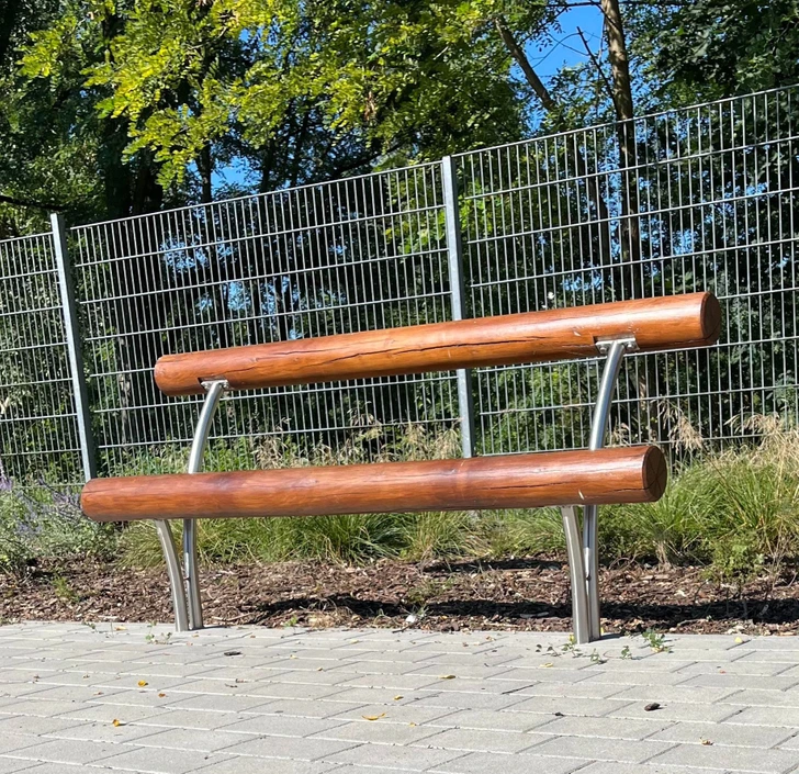 Moderná drevená lavička s kovovou konštrukciou umiestnená na dlaždenom chodníku v parku.