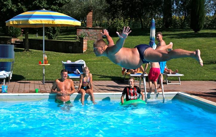 Muž skáče šípku do bazéna, ľudia sledujú s úsmevom. Slnečný deň pri bazéne plnom zábavy.