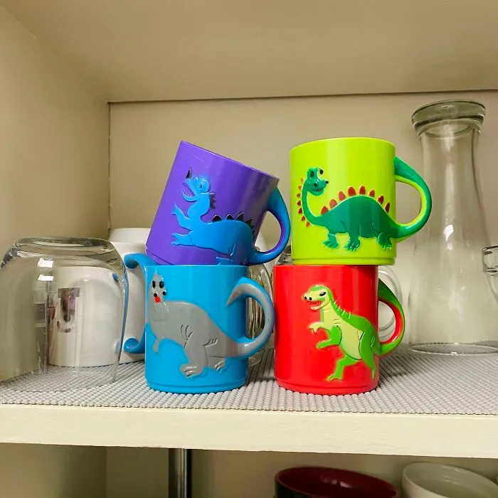 Farebné hrnčeky s dinosaurami v poličke, usporiadané a pripravené na použitie v detskej kuchyni.