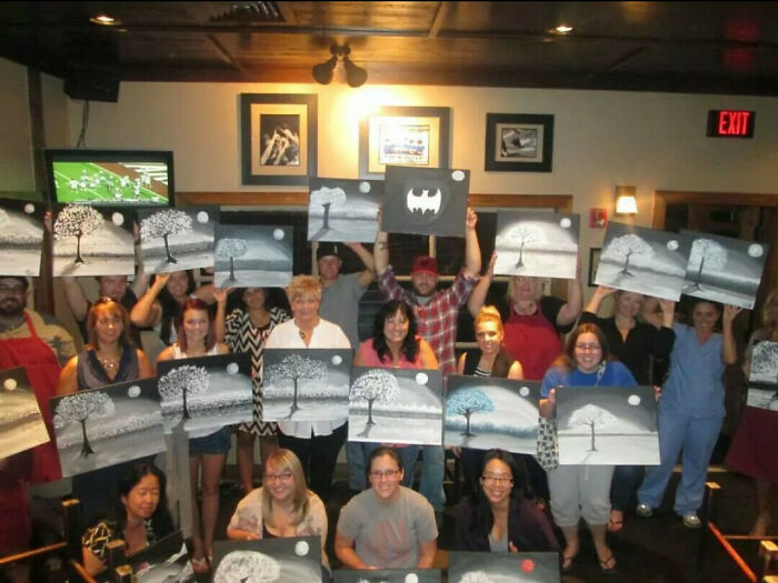 Skupina ľudí v interiéri drží svoje obrazy s motívom stromu na spoločnom umeleckom podujatí.