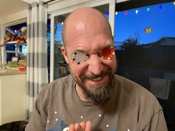 Muž s pokerovými žetónmi na očiach sa smeje v obývačke s rozsvietenými svetielkami.