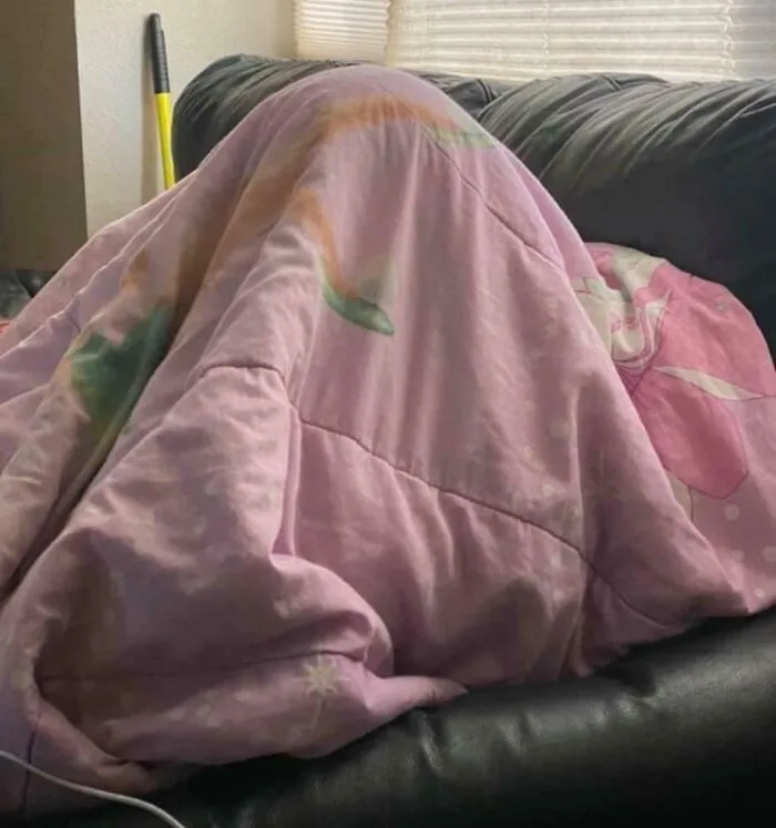 Osoba ukrytá pod ružovou dekou na pohovke, vytvára pohodlný a tajomný úkryt v obývačke.