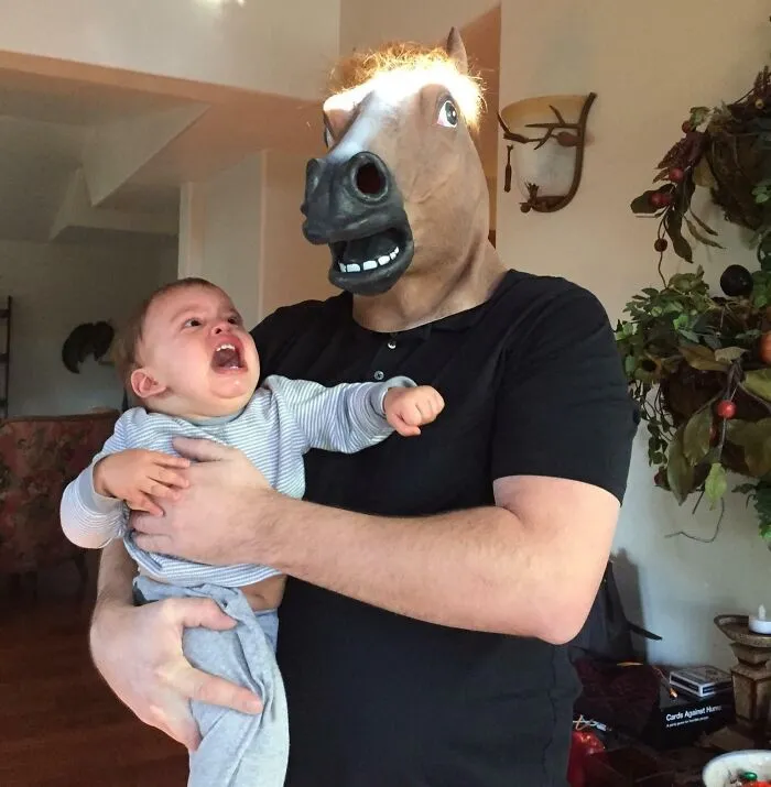 Osoba s maskou koňa drží plačúce dieťa v obývačke, podivná a vtipná situácia.